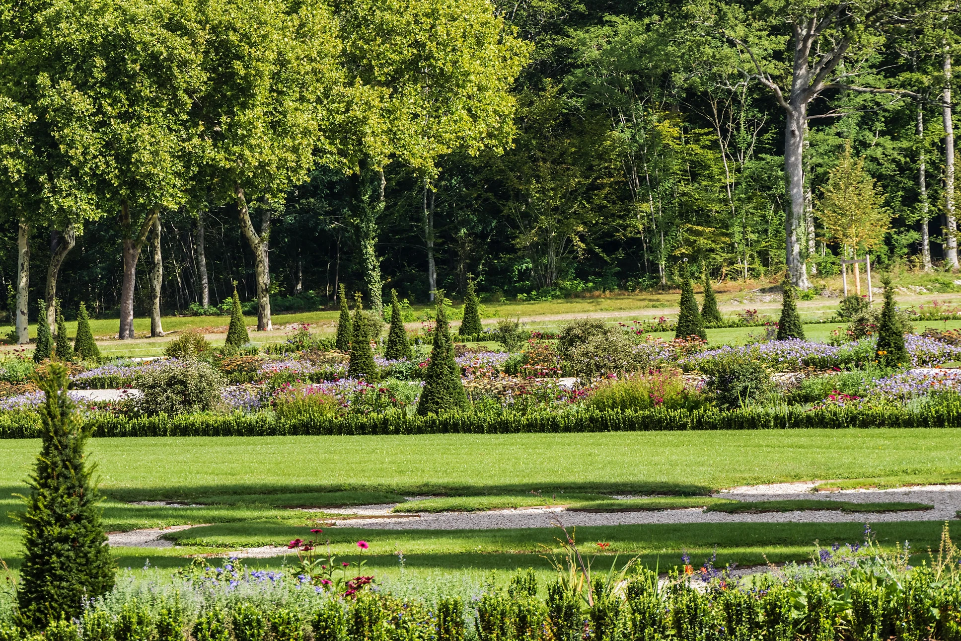 Jardin paysager avec pelouse entretenue, massifs floraux colorés délimités par bordures buis taillées, topiaires coniques, allées gravillonnées